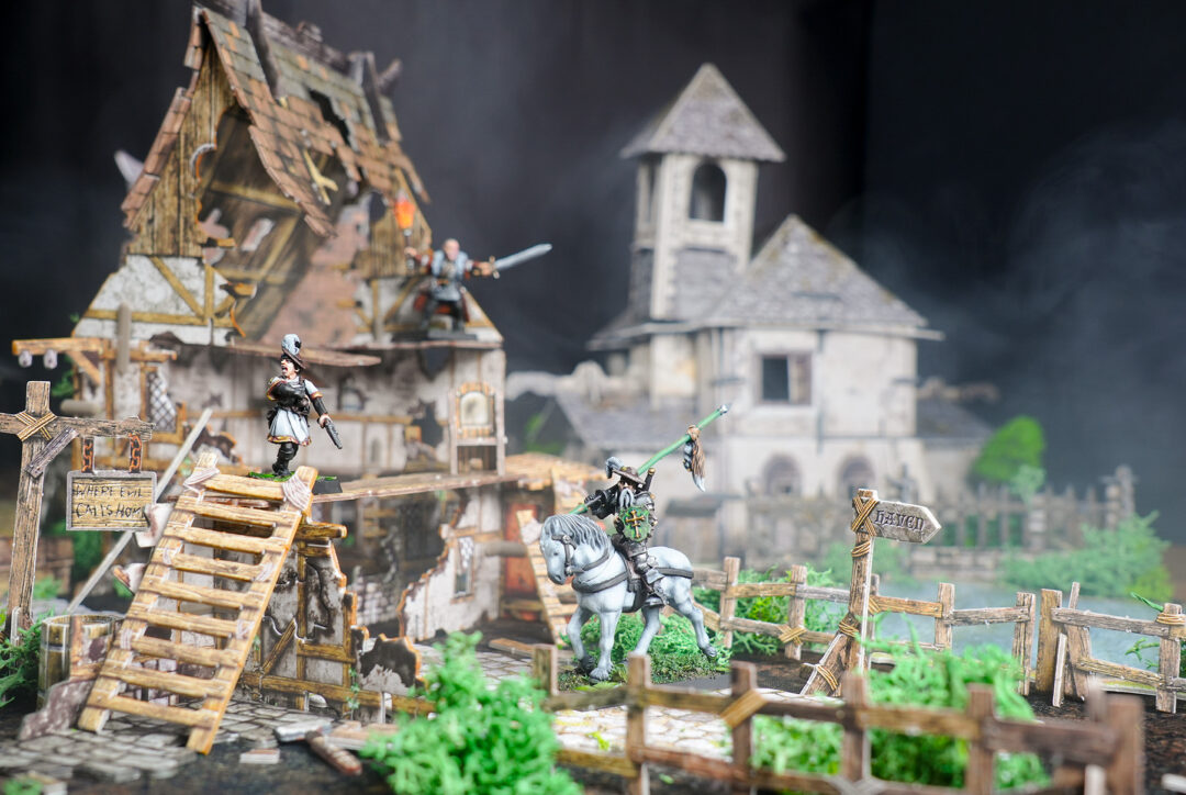 A warband defends multilevel Mordheim terrain including a ruined house, a chapel stands in the background.
